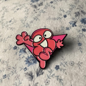 Pu&ograve; includere: Spilla in smalto rosa raffigurante un personaggio dei cartoni animati con un volto sorridente, occhi bianchi e uno sfondo triangolare rosa. La spilla ha un contorno nero, su uno sfondo a motivi floreali.