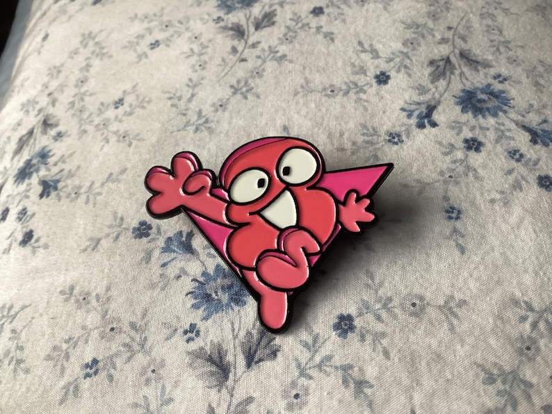 Pu&ograve; includere: Spilla in smalto rosa raffigurante un personaggio dei cartoni animati con un volto sorridente, occhi bianchi e uno sfondo triangolare rosa. La spilla ha un contorno nero, su uno sfondo a motivi floreali.
