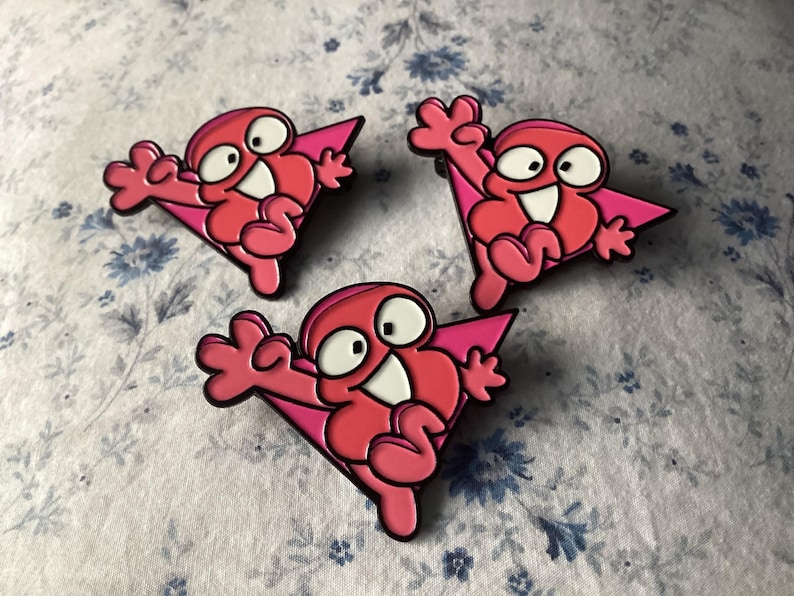 Pu&ograve; includere: Tre spille smaltate rosa raffiguranti un personaggio dei cartoni animati con un viso bianco, grandi occhi e un ampio sorriso. Il personaggio indossa un mantello rosa. Le spille sono su un tessuto a fantasia floreale bianco e blu.
