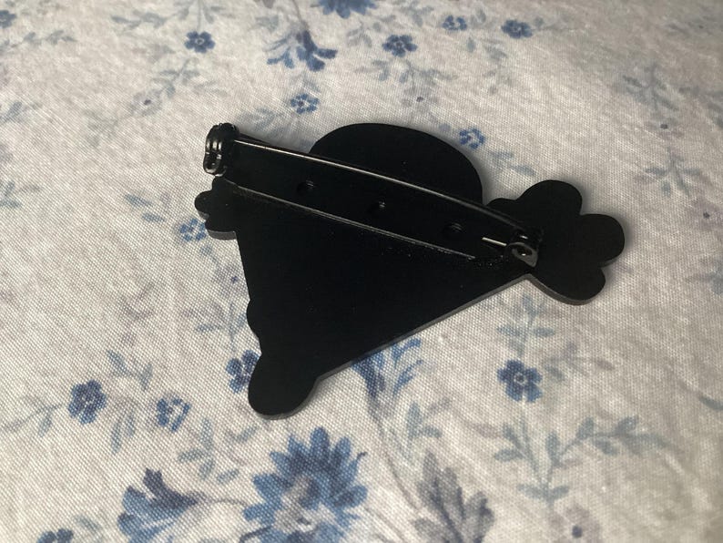 Pu&ograve; includere: Una spilla nera dal design unico, con una parte superiore arrotondata e una forma stilizzata con un elemento a forma di fiore. La spilla ha una chiusura di sicurezza sul retro per un fissaggio sicuro. Questo &egrave; probabilmente un accessorio di moda.