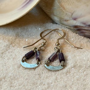May include: A pair of dangle earrings with a silver-tone metal crescent and dark purple and white beads. The earrings are set against a backdrop of sand and seashells, creating a beachy aesthetic.