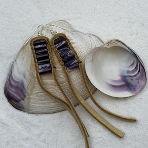 May include: Dangle earrings with silver hooks. Each earring has a rectangular, dark purple and white striped bead design, framed in brown leather and attached to long, tan leather strips. The earrings are displayed on a seashell.