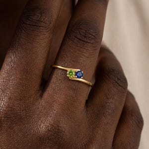 May include: A gold bypass ring featuring two round-cut gemstones, one vibrant green peridot and one deep blue sapphire, set in a delicate polished gold band that overlaps slightly at the centre to hold the two stones securely in place.