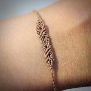Puede incluir: Una pulsera de macramé de color marrón claro con una sección central de intrincados patrones de nudos repetitivos. El cordón fino está tejido en una serie de pequeños bucles en forma de diamante que crean un diseño continuo y texturizado.