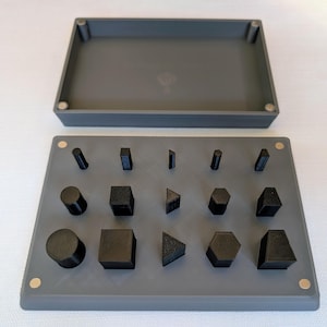 May include: A gray, rectangular tray with a matching lid, containing various black geometric shapes. The shapes include cylinders, cubes, triangles, and hexagons. The tray has small magnets in each corner.