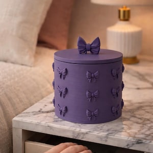 May include: A lilac decorative box with a bow-shaped handle on the lid and bow accents around the sides. The box is on a marble-topped bedside table next to a lamp.