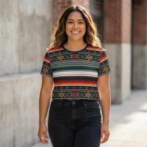 May include: A black crop top with a Southwestern-inspired pattern. The design features geometric shapes in red, yellow, green, and white, with horizontal stripes in various colors. The shirt has short sleeves and a round neckline.