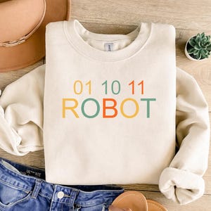 May include: A cream-colored sweatshirt with the word "ROBOT" in orange, green, and yellow letters. Above the word are the numbers "01 10 11" in the same colors. The sweatshirt is folded on a light-colored wooden surface.