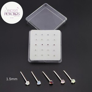 Puede incluir: Una caja de plástico transparente que contiene una selección de piercings de nariz con piedras preciosas de varios colores. Se muestran cinco piercings individuales con gemas de 1,5 mm debajo de la caja. El texto "Nose Piercings" está en un círculo en la esquina superior izquierda.