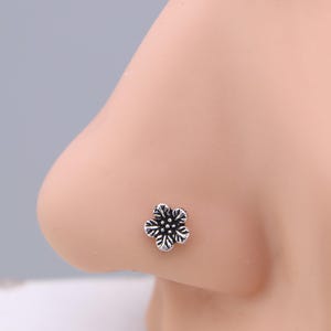 May include: A silver nose stud shaped like a five-petal flower. The floral design features detailed lines and a slightly oxidized finish. The stud is set against a light skin tone.