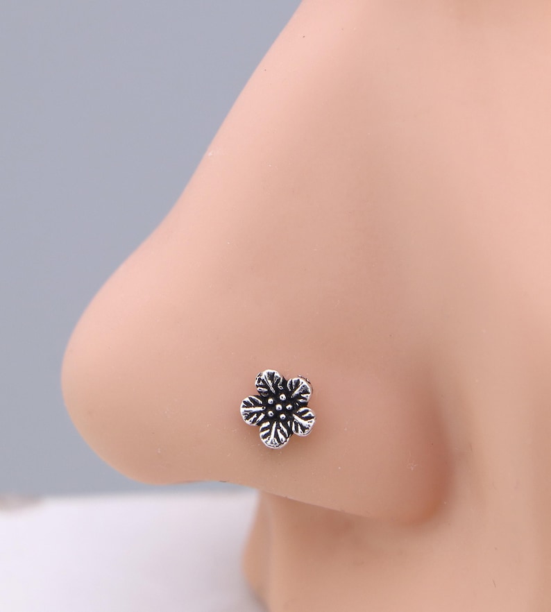 May include: A silver nose stud shaped like a five-petal flower. The floral design features detailed lines and a slightly oxidized finish. The stud is set against a light skin tone.