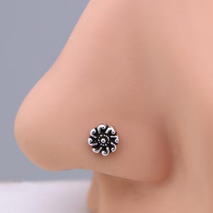 May include: A silver nose stud shaped like a flower. The floral design has detailed petals and a central accent. The jewellery is shown on a person's nose, highlighting the stud's size and design.