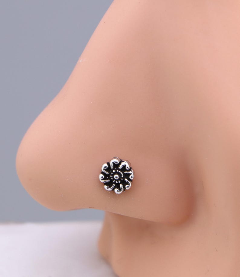 May include: A silver nose stud shaped like a flower. The floral design has detailed petals and a central accent. The jewellery is shown on a person's nose, highlighting the stud's size and design.