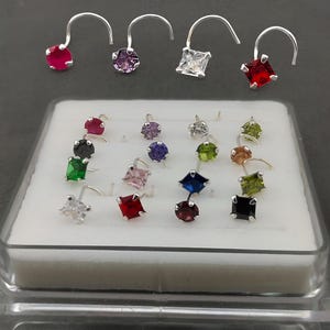 May include: A collection of nose rings with various colored gemstones, including red, purple, green, and clear, set in silver-toned metal. The gemstones are round, square, and diamond-shaped. The nose rings are displayed in a white tray.