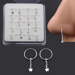 Puede incluir: Anillos de nariz plateados con colgantes de estrella. Se muestran en una caja de plástico transparente. Un primer plano muestra una nariz con uno de ellos. Se muestran dos anillos adicionales.