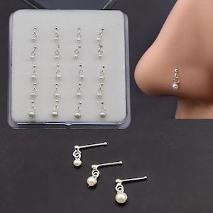 Könnte beinhalten: Eine Sammlung silberner Nasenpiercings mit baumelnden weißen Perlen. Die Nasenpiercings werden in einer transparenten Kunststoffbox präsentiert und auch an der Nase eines Models gezeigt. Drei zusätzliche Nasenpiercings werden separat gezeigt.