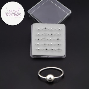 Può includere: Un set di piercing al naso argentati con una piccola sfera al centro, esposti in una custodia di plastica trasparente. L'immagine mostra anche un singolo piercing al naso con una sfera. Il logo "Nose Piercings" è visibile.