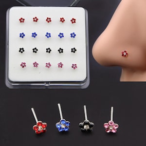 May include: A set of flower-shaped nose studs in a variety of colors, including red, blue, black, and pink. Each stud features a central clear gem. The studs are displayed in a clear plastic case.