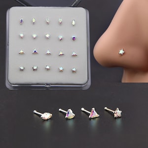 May include: A set of nose studs in various shapes, including teardrops, hearts, triangles, and stars, with iridescent, multi-colored crystals. The studs are displayed in a clear plastic case and individually. One star-shaped stud is shown on a nose.