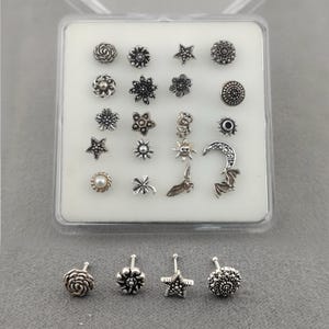 以下が含まれることがあります： 透明なプラスチックケースに入った、銀色のノーズスタッドのセット。スタッドは、花、星、三日月、コウモリなど、さまざまなデザインが特徴です。スタッドは白い表面に表示されています。