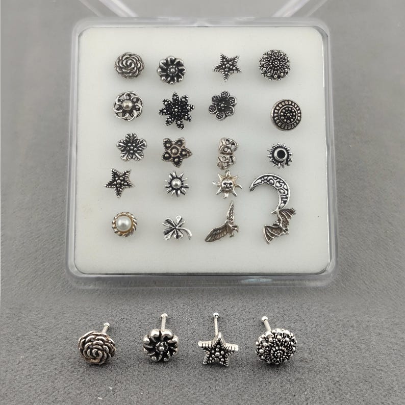 May include: A collection of silver-coloured nose studs presented in a clear plastic case. The studs showcase diverse designs, such as flowers, stars, a crescent moon, and a bat. The studs are arranged on a white surface.