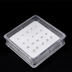May include: A clear plastic box containing multiple small, round, silver-coloured nose studs. The studs are set on a white, cushioned surface. The box is square and has a transparent lid, set against a black background.
