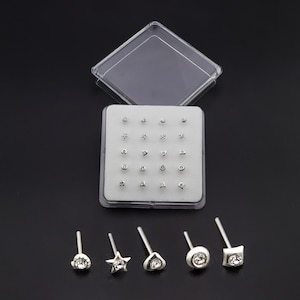 Puede incluir: Un conjunto de piercings para la nariz de color plateado en una caja de plástico transparente. Los piercings presentan varias formas, incluyendo estrellas, círculos y cuadrados, cada uno con una pequeña gema transparente. La caja está abierta, revelando la disposición de los piercings.