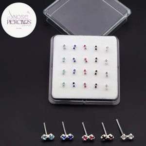 May include: A set of nose piercings in a clear plastic case. The piercings are small, with a stud design featuring a cluster of colorful gemstones in various colors, including blue, red, and clear. The text "Nose Piercings" is visible in the upper left corner.