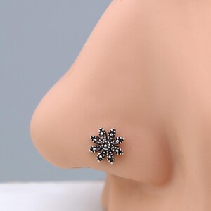 May include: A silver-coloured nose stud shaped like a flower. The floral design has multiple small, round petals around a central cluster. The jewellery is set against a light skin tone.