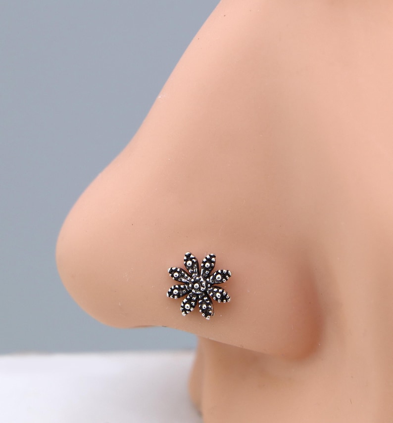 May include: A silver-coloured nose stud shaped like a flower. The floral design has multiple small, round petals around a central cluster. The jewellery is set against a light skin tone.