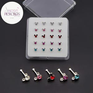 May include: A set of nose piercings in a clear plastic case. The piercings are shaped like a mouse with a crystal or colored gem. Colors include clear, pink, red, purple, and blue. The text "Nose Piercings" is in a white circle.