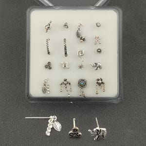 May include: A collection of silver-toned nose studs in various designs, including leaves, spirals, and an elephant. The studs are displayed in a clear plastic case, with three additional studs visible below. The designs are intricate and detailed.