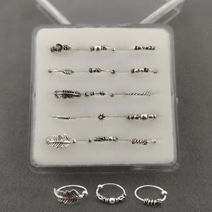 Puede incluir: Una colección de piercings de nariz plateados, exhibidos en una caja de plástico transparente. Los anillos presentan varios diseños, incluyendo plumas, cuentas y alambre retorcido. La caja está sobre una superficie gris.