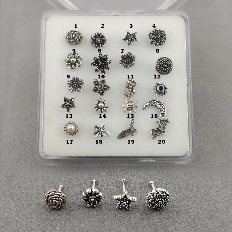 May include: A selection of twenty silver-coloured nose studs in diverse designs, such as floral, star, and geometric patterns. The studs are presented in a clear plastic case, with four studs visible below the case.