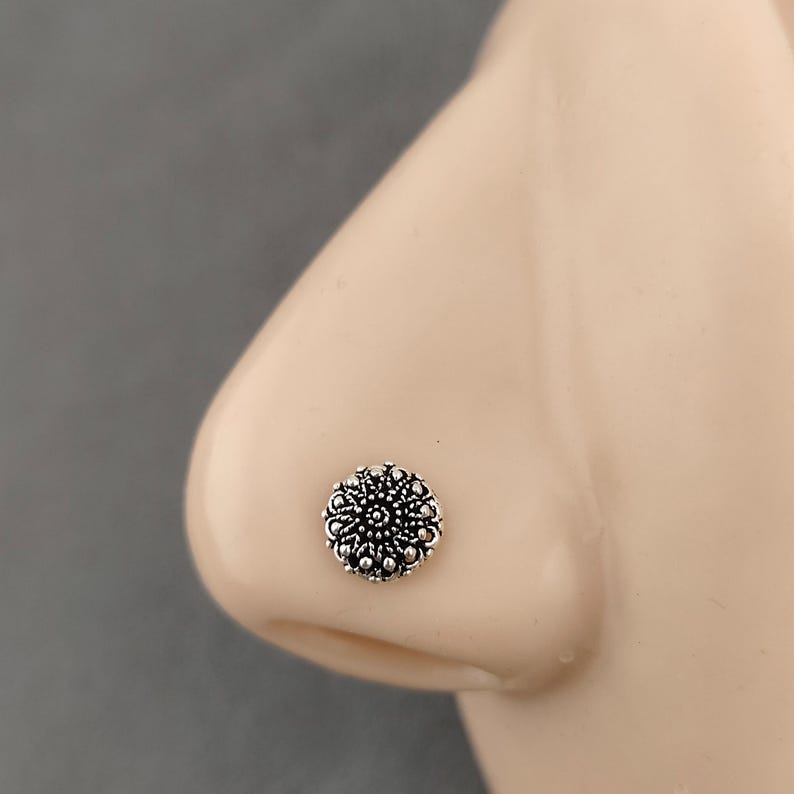 May include: A silver nose stud with a floral pattern. The stud is circular and has a detailed design of small, raised dots and curved lines, giving it a textured look. The jewellery is set against a neutral skin tone background.