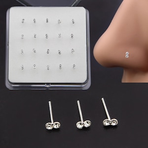 May include: A set of silver-toned nose studs featuring two small, clear, round crystals arranged in a vertical line. The collection is displayed in a square white foam insert within a clear plastic case, with three individual studs placed below.
