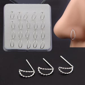 May include: A collection of silver-colored nose rings in various styles. The image shows a tray of oval-shaped rings with a twisted design, a nose with a teardrop-shaped ring, and three half-moon rings with a twisted design.