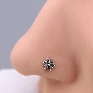 Könnte beinhalten: Ein silberner Nasenstecker in Form einer Blume. Der kleine Stecker hat ein detailliertes Design mit einer zentralen Perle und umliegenden Blütenblättern. Der Schmuck befindet sich auf einem hellen Hautton.