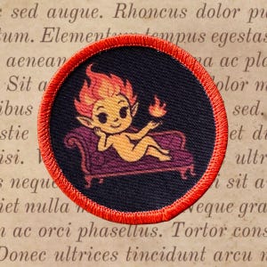 May include: Round patch with a red stitched border. The patch features a cartoon imp with orange hair and skin, reclining on a purple couch. The imp holds a small flame in its hand against a dark blue background.
