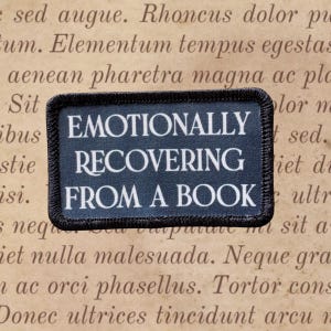May include: A rectangular patch with the words "EMOTIONALLY RECOVERING FROM A BOOK" in white text on a dark blue background. The patch has a black border and is set against a backdrop of aged paper with faded text.