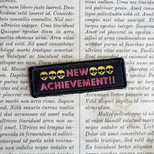 May include: A rectangular black embroidered patch featuring the yellow text NEW and the pink text ACHIEVEMENT!!, flanked by three yellow skull icons on each side. The patch has a black stitched border and is set against a background of Latin text.