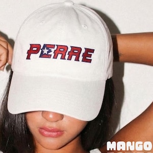 May include: White baseball cap with the word "PERRE" embroidered in red and blue. The cap features a curved brim and a classic six-panel design. The text is in a bold, stylized font. The cap is a casual accessory.
