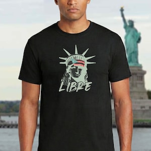 May include: Black t-shirt featuring a distressed graphic of the Statue of Liberty with a Puerto Rican flag in the crown and the word "LIBRE" in white. The background shows the Statue of Liberty.