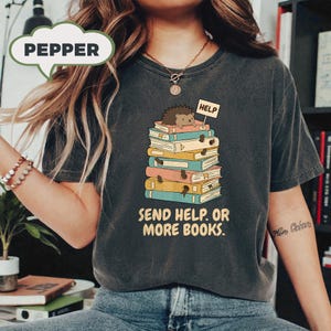 Puede incluir: Camiseta gris oscuro con un diseño de un erizo sobre una pila de libros coloridos. El erizo sostiene un cartel de "HELP". El texto "SEND HELP. OR MORE BOOKS." está debajo. La camiseta tiene un aspecto vintage.