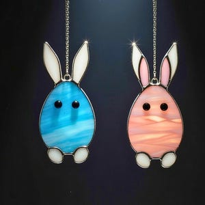 Könnte beinhalten: Zwei Osterhasen-Ornamente. Eines ist blau mit weißen Ohren und Füßen, das andere ist rosa mit weißen Ohren und Füßen. Beide haben schwarze Augen und hängen an silbernen Ketten. Die Ornamente sind aus Glas gefertigt.