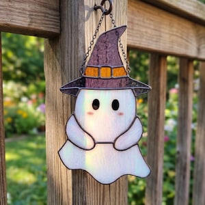 Può includere: Un acchiappasole in vetro colorato a forma di fantasma che indossa un cappello da strega viola con una fascia arancione. Il fantasma è realizzato in vetro bianco iridescente con due occhi neri circolari e piccoli accenti rosa sulle guance, sospeso da una catena.