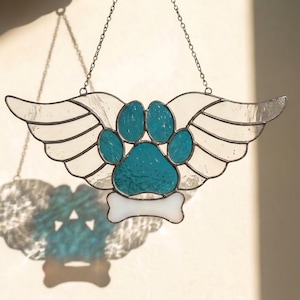 May include: Stained glass suncatcher featuring a turquoise paw print with white wings and a bone. The suncatcher is suspended by a chain. The wings and bone are white, and the paw print is a textured turquoise color.