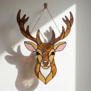 May include: Stained glass deer head with brown, tan, and pink hues. The deer has large antlers and is suspended by a chain. The artwork is designed to be hung on a wall and casts a shadow.