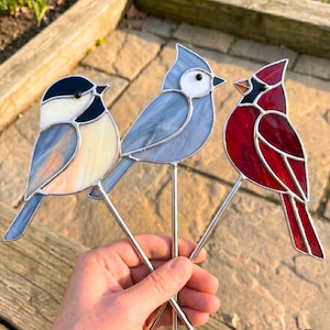 Op de afbeelding: Drie glas-in-lood vogelornamenten op metalen staven. De linker vogel is crème en zwart met blauwe accenten. De middelste vogel is lichtblauw met een wit gezicht. De rechter vogel is dieprood met een zwart gezicht en een oranje snavel.