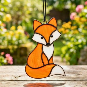 May include: A stained glass fox ornament with orange, white, and black details. The fox is suspended by a black cord. The ornament is set against a blurred background of greenery and flowers.